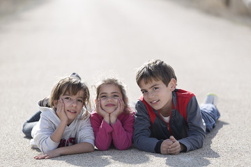 Three children lying on the ground of a road facing the camera.