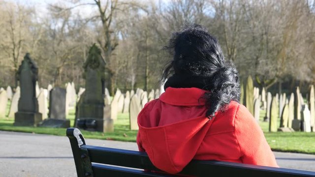 Lone sad dark long hair woman in red coat is sitting on bench in an old English cemetery on a sunny windy day in autumn or winter, rear view
