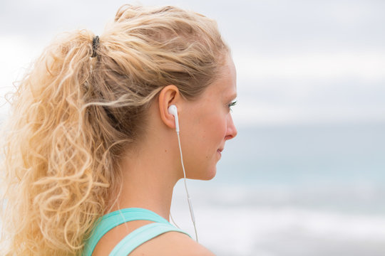 Fitness girl listening to music with sport earphones. Caucasian blonde female athlete blonde woman runner wearing earbuds ready to go run on beach, summer active lifestyle. Portrait closeup of ears.