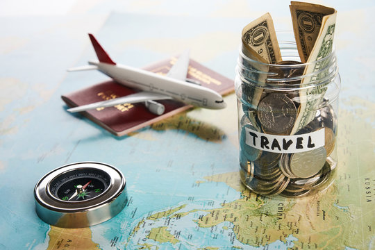 Holidays money savings in a glass jar with compass, passport travel document and plane toy on world map, close-up. Travel budget concept.