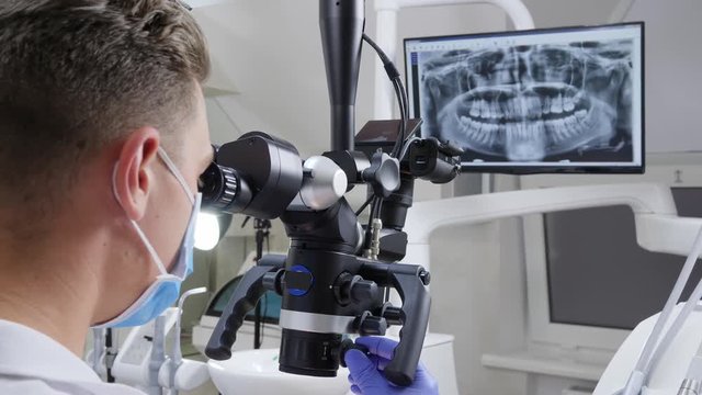 dentist in medical mask adjusts binocular microscope to work, roentgen of jaw on screen on background