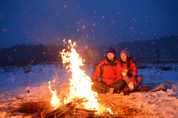 Happy loving couple sitting at a fire in the snow among the fields in the winter against the background of the forest and the sky on Valentine's Day.