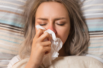 Young European upset and sick female with paper napkin blowing nose, lies on the pillow, closed eyes, indoor/ Sinusitis treatment / Rhinitis, cold, sickness, allergy concept/