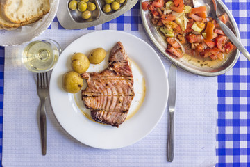 Tuna steak accompanied with potatoes, olives, tomato salad, bread and wine