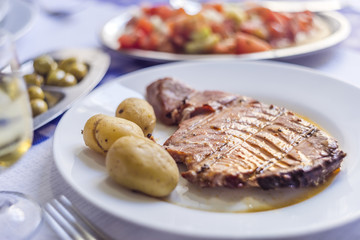 Tuna steak accompanied with potatoes, olives, tomato salad and wine