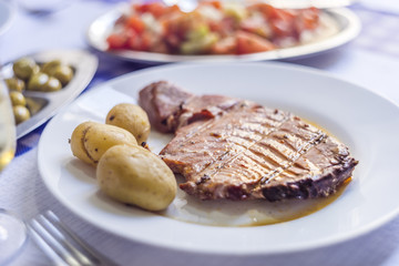 Tuna steak accompanied with potatoes, olives, tomato salad and wine