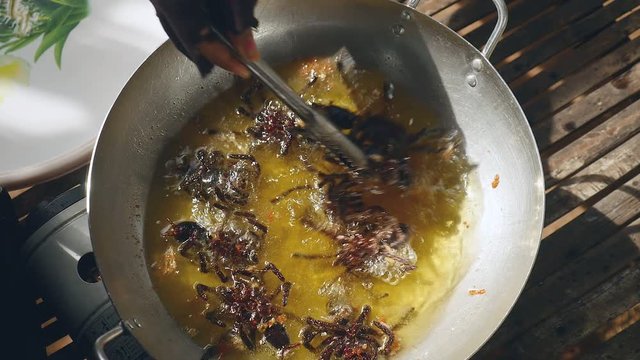 Woman turning over deep frying tarantulas inside a wok using metal tongs (close-up upper view)