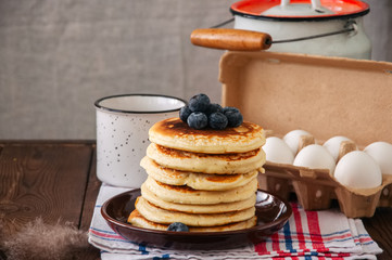 Stack of homemade pancakes with blueberries in a plate, milk in a flask, eggs in a paper container. Rustic style, still life. Copy space.