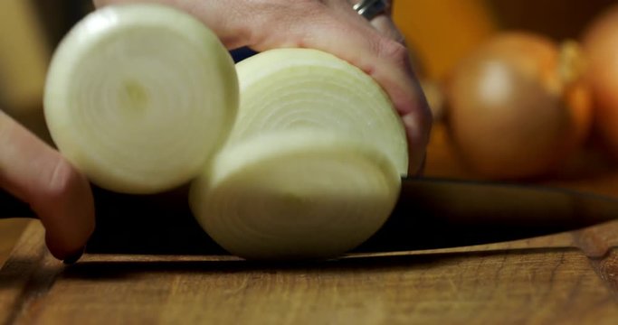 Woman's hand holding chopping knife and slicing an onion on wooden board in kitchen. Recorded in 4K with dolly move.