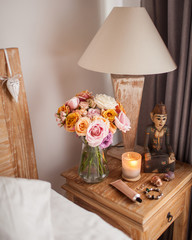 Wooden Bed with White Sheets. A Bedside Table by the Bed with a Lamp, a Bouquet of Flowers from Roses, Candle and Hand Cream