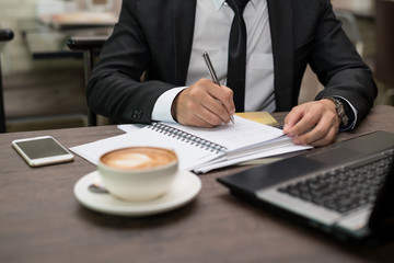 Young Asian businessman writing notes sitting in a coffee shop. On the table is a laptop, a smart phone and a cup of coffee.