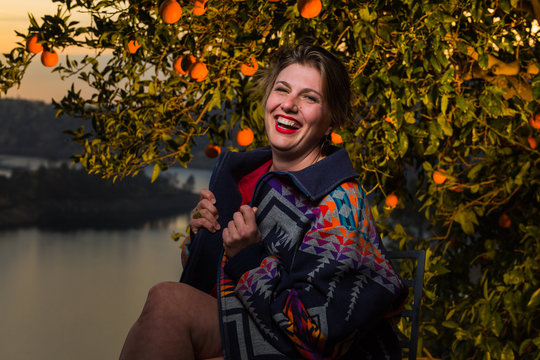Young caucasian woman with short hair and flashy jacket poses under an orange tree at sunset