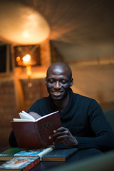 A young Arican man is reading a book in a room at a table. student, businessman