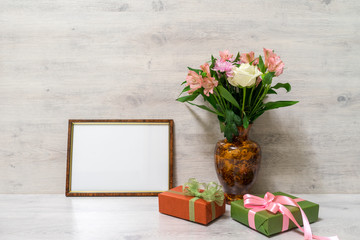 Colorful spring bouquet of rose, chrysanthemum and alstroemeria flowers in a vase with empty photoframe and gift boxes on wooden background
