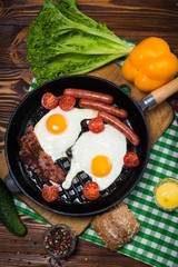 Fried eggs with bacon and sausages in a frying pan with spices and fresh vegetables.