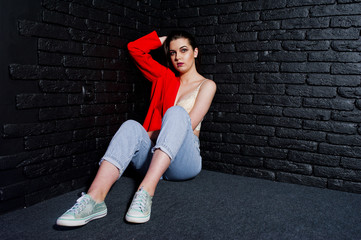 Stylish brunette girl on red jacket against studio black brick wall.