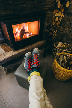 POV photo from above of man resting his feet in hard labor leather winter boots in front of warm and cosy hot fireplace, burning in living room or lobby of hotel in ski mountain resort
