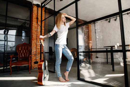 Portrait of the stylish young woman of the blonde in a white t-shirt and jeans in an interior in style the loft. The girl with a guitar.
