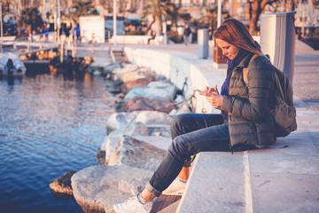 young woman with mobile phone in hands sits on the waterfront in a beautiful European city at sunset