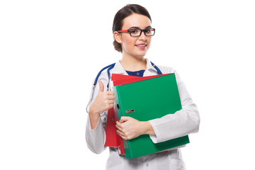Young woman doctor with stethoscope holding binders in her hands with thumb up in white uniform on white background