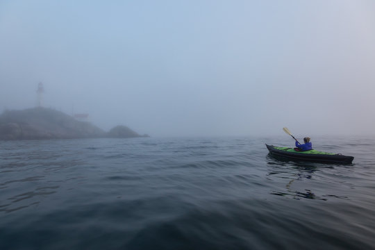Adventurous woman is sea kayaking near a lighthouse during a vibrant and foggy winter sunset. Taken in Horseshoe Bay, West Vancouver, BC, Canada. Concept: adventure, holiday, vacation, explore