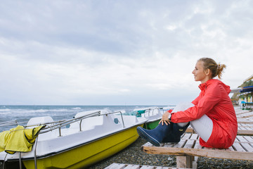 young woman in a red jacket and blue rubber boots sitting on a wooden deck chair and looking at the stormy sea