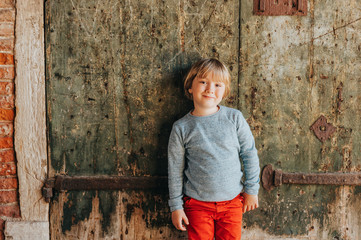 Outdoor portrait of handsome little boy wearing red chinos and light blue pullover, stylish kid posing against vintage old wall. Fashion for small children