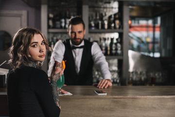 bartender and tattooed girl looking at camera at bar counter