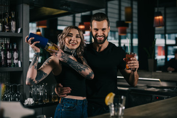 smiling young bartenders preparing alcohol drinks at bar and looking at camera