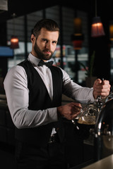 handsome bartender pouring beer from beer taps into glass and looking at camera