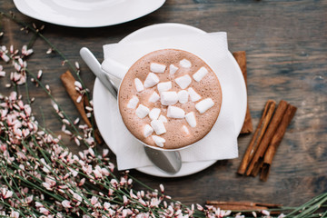 Delicious hot chocolate with marshmallows on top with cinnamon and little pink flowers on the background, rustic wooden table