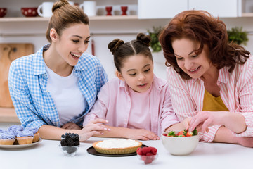 three generations of women decorating dessert with berries together at kitchen