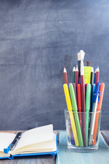 Glass with pencils and notepad on a black chalkboard, wooden table