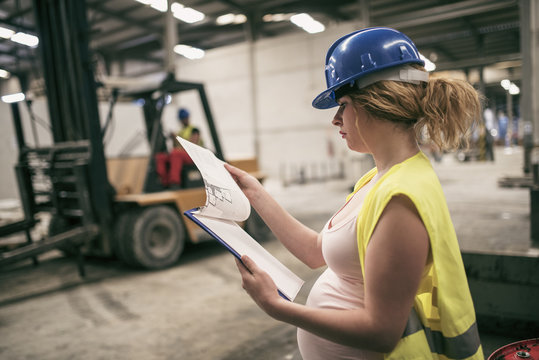 Pregnant woman working in construction factory. Man driving forklift in background