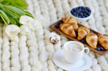Obraz na płótnie Canvas Cup with cappuccino and croissants, berries, white pastel giant knit blanket, bedroom, flowers tulips, spring, woman day, morning concept 