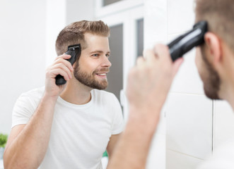 Handsome man cutting his own hair with a clipper - obrazy, fototapety, plakaty