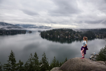 Fototapeta na wymiar Young Caucasian Girl is enjoying a beautiful scenery from the top of a mountain during a cloudy winter morning. Taken in Quarry Rock, Deep Cove, North Vancouver, BC, Canada.