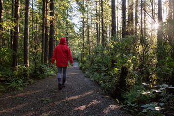 Fototapeta premium Girl wearing a bright red jacket is walking the the beautiful woods during a vibrant winter morning. Taken in Ucluelet, Vancouver Island, BC, Canada.