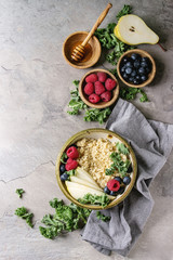 Vegan quinoa porridge with kale, strawberries, blueberries, sliced pear, honey on dipper in bowl with ingredients above and cloth napkin over grey texture background. Top view, space. Healthy eating