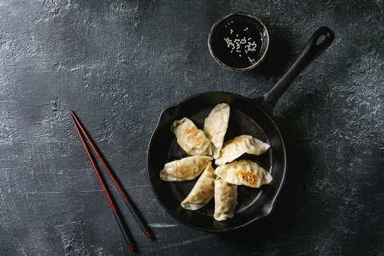 Asian dumplings Gyozas potstickers fried on cast-iron pan, served with chopsticks and bowl of soy sesame sauce over black texture background. Top view, space.