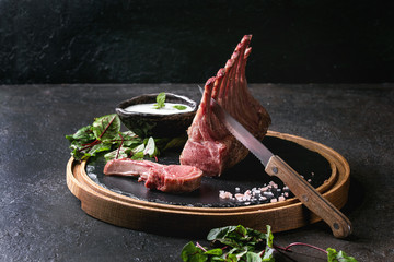 Grilled sliced rack of lamb with yogurt mint sauce served with green salad young beetroot leaves, knife and pink salt on round wooden slate board over dark black table background. Copy space © Natasha Breen