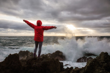 Woman in a bright red jacket is watching waves crash on a rocky Pacific Ocean Coast. Taken in Ucluelet, Vancouver Island, BC, Canada.