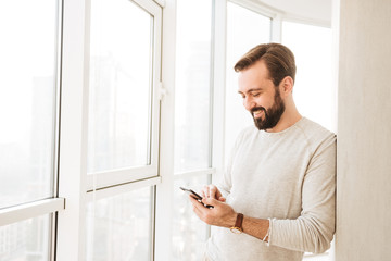 Photo of relaxed guy 30s having beard and mustache, spending time in flat and using mobile phone near big window