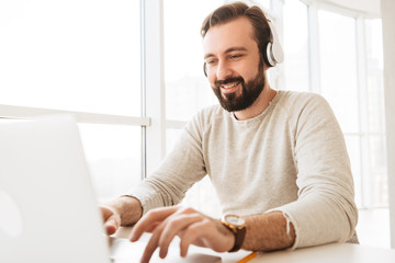 Photo of attractive mature man 30s in casual clothing typing text message or working on laptop, while listening to music via earphones