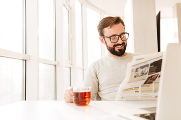 Handsome bearded man wearing glasses drinking tea from glass and reading newspaper, while having...