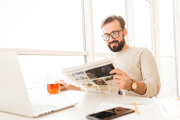 Brunette bearded man wearing eyeglasses drinking tea from glass and reading newspaper, while resting at home