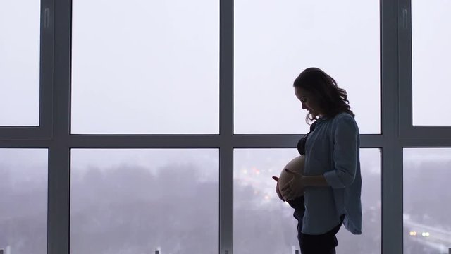 A happy pregnant woman stands at a large panoramic window located at a high altitude. He touches his stomach with his hands and communicates smilingly with the baby in the stomach. He looks at the