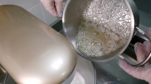 Boiling sugar liquor is being poured to the electric beater for cake producing. Female confectioner is holding steel pot by her hands in latex gloves and additing small portions of syrop to the white