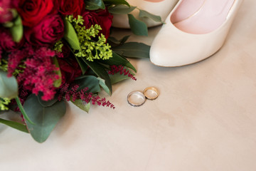 Obraz na płótnie Canvas Golden wedding rings in focus on beige background with bridal shoes and flowers out of focus.