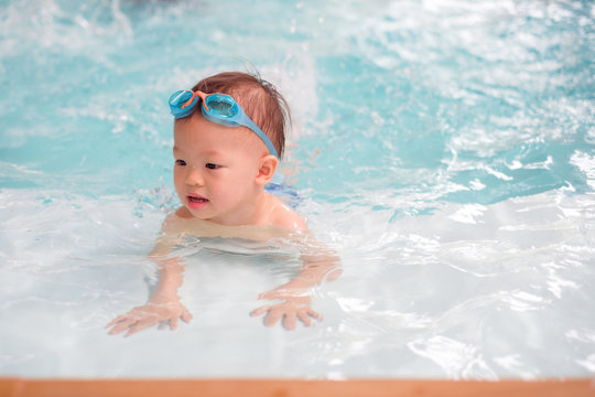 Cute little Asian 18 months / 1 year old toddler boy child in trunks wear swimming goggles learn to swim at indoor pool, Swimming school for small children. Happy kid enjoy active lifestyle concept